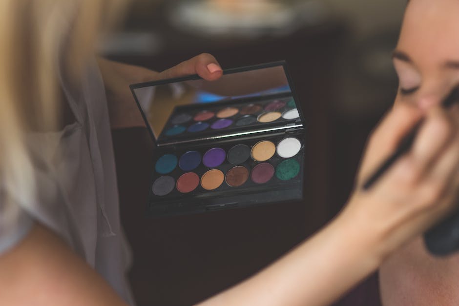 A close-up of a stylist applying eyeshadow from a vibrant color palette to a model's face indoors
