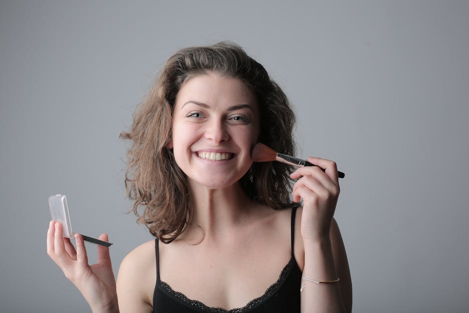 A joyful woman applies makeup with a brush, posing in a studio with a bright smile